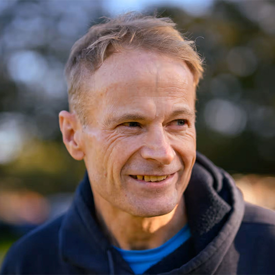 A man in a hooded jumper smiling and looking sidewards away from the camera.